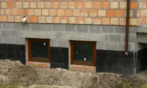 Ocieplanie fundamentów starego domu: widok na fragment ściany z oknami piwnicznymi, izolacją przeciwwilgociową i warstwą ocieplenia.