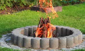 Ognisko na działce w betonowej obudowie, płonące drewno na tle zielonej trawy. Czy można palić ognisko na działce? Tak, jeśli jest bezpiecznie!