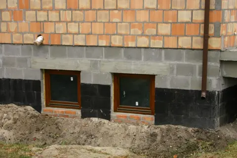 Ocieplanie fundamentów starego domu: widok na fragment ściany z oknami piwnicznymi, izolacją przeciwwilgociową i warstwą ocieplenia.