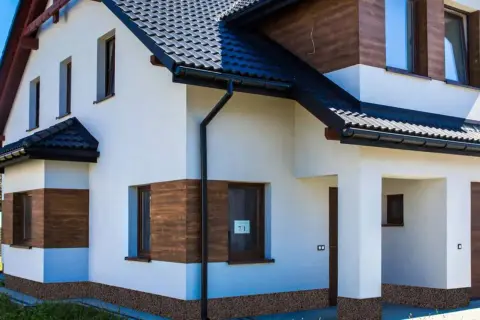 Nowy dom z białym tynkiem i drewnianymi akcentami. Brak cokołu na elewacji, widoczna kamyczkowa podmurówka.