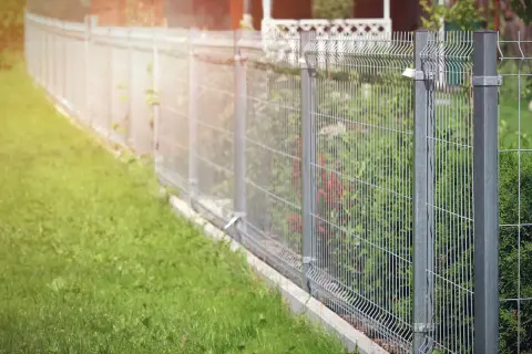 Solidne, metalowe ogrodzenie panelowe to świetny pomysł na tanie ogrodzenie działki. Widać zieloną trawę i krzewy za siatką.