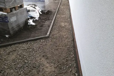 Wykonano opaskę wokół domu z płyt chodnikowych, otaczającą żwirowany teren budowy z paletami kostki brukowej.