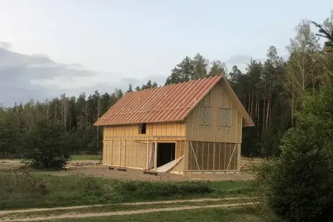 Trwa budowa domu ze słomy. Widać drewnianą konstrukcję, dach i ściany z widocznymi belami słomy.