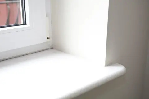 Biały, nowy parapet wewnętrzny po wymianie, z zaokrąglonym rogiem, gotowy na nowe dekoracje.