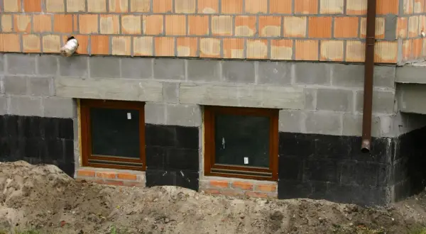 Ocieplanie fundamentów starego domu: widok na fragment ściany z oknami piwnicznymi, izolacją przeciwwilgociową i warstwą ocieplenia.