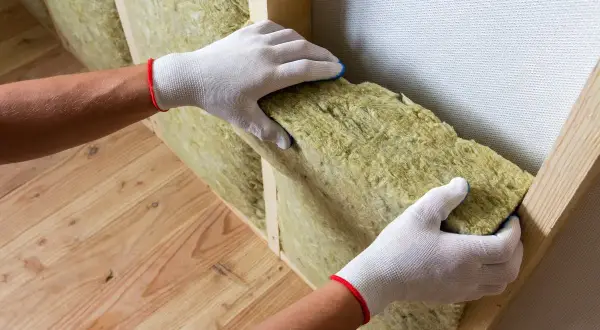 Ręce w białych rękawiczkach montują wełnę mineralną. To dobry sposób, czym ocieplić ściany od wewnątrz, by było ciepło i cicho.