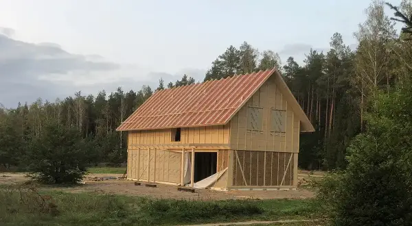Trwa budowa domu ze słomy. Widać drewnianą konstrukcję, dach i ściany z widocznymi belami słomy.