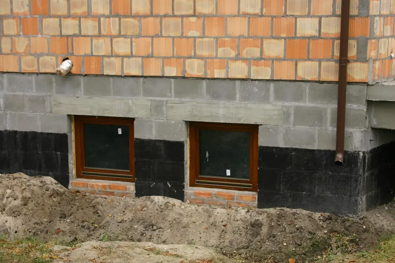 Ocieplanie fundamentów starego domu: widok na fragment ściany z oknami piwnicznymi, izolacją przeciwwilgociową i warstwą ocieplenia.