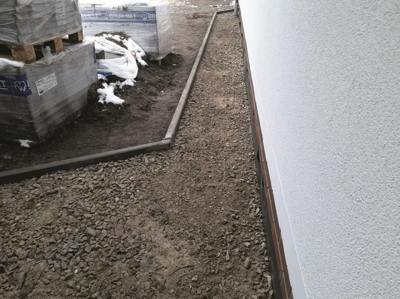 Wykonano opaskę wokół domu z płyt chodnikowych, otaczającą żwirowany teren budowy z paletami kostki brukowej.