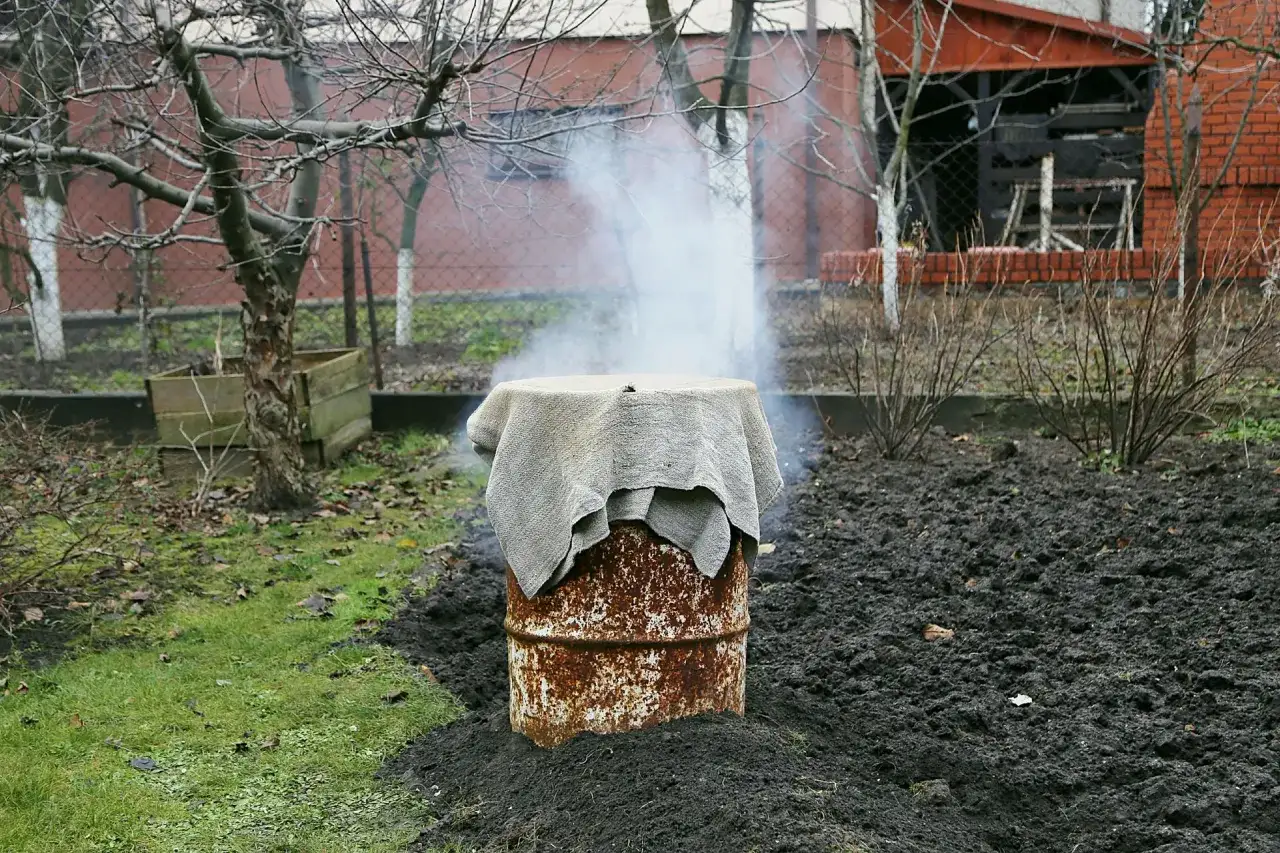Dym unosi się z beczki, która służy jako prowizoryczna wędzarnia. Wokół niej ziemia i trawa.