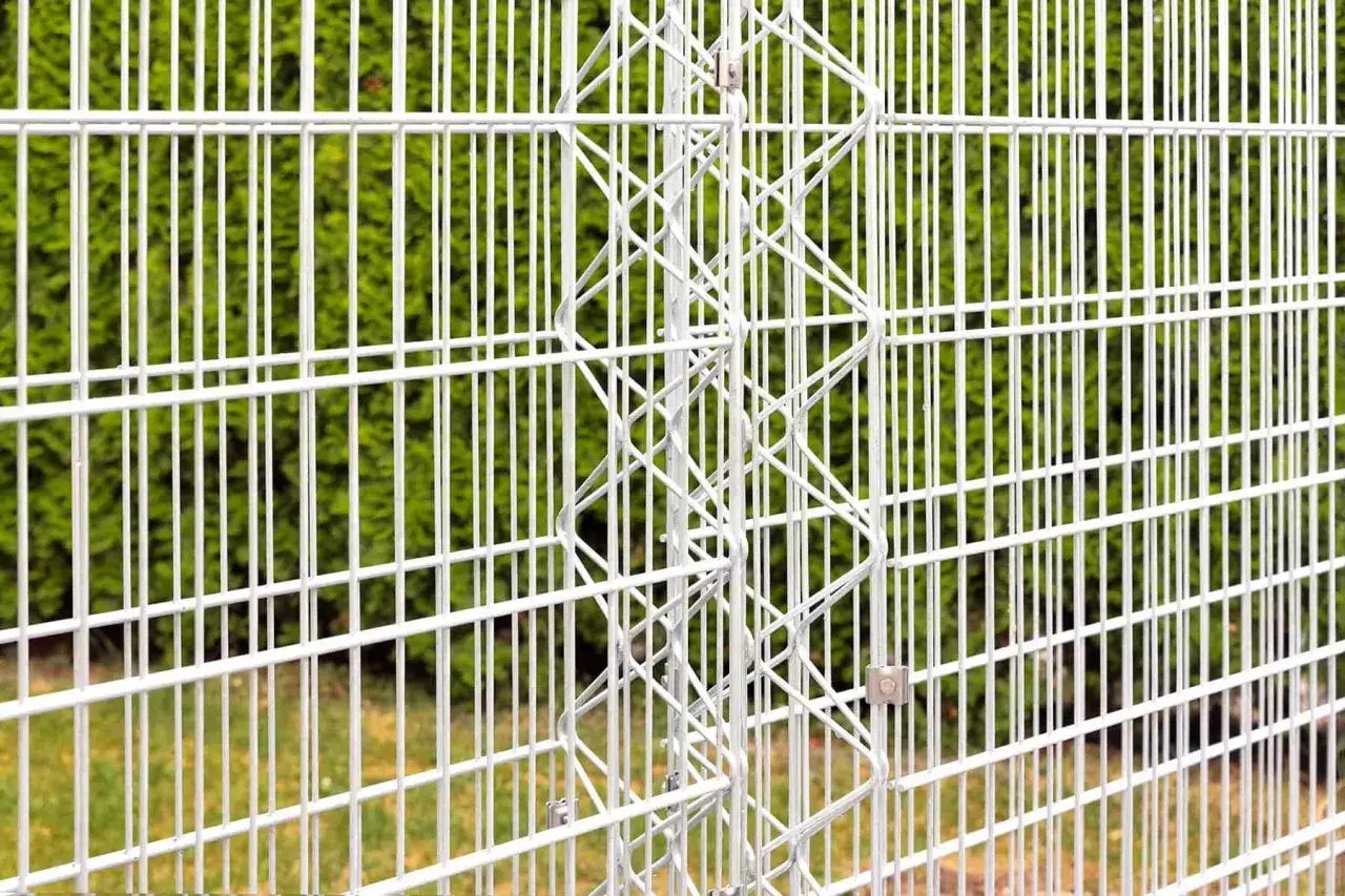 Białe, metalowe ogrodzenie posesji z ozdobnym, spiralnym elementem. Tło stanowi zielony żywopłot.