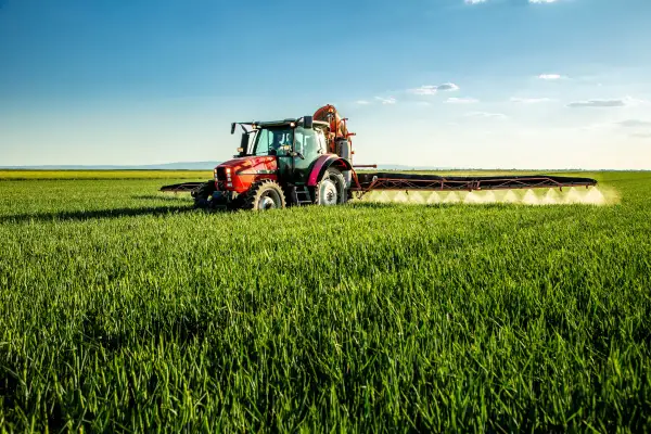 Czerwony traktor wykonuje oprysk na dwuliścienne na zielonym polu pod błękitnym niebem.